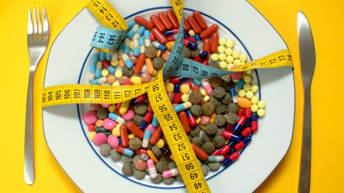 Auf einem Teller liegen viele bunte Pillen, ein Maßband ist kreuzweise über den Teller gebunden.