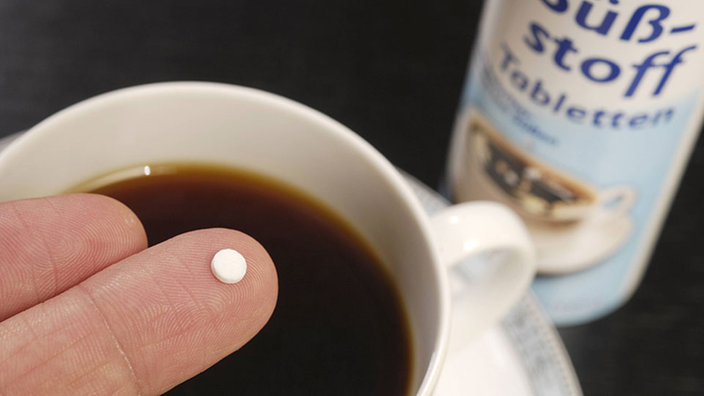 Mit Kaffee gefüllte Porzellantasse, darüber hält eine Hand einen Süßstoffspender aus dem eine kleine Tablette fällt.