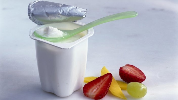 Ein weißer Becher mit Naturjoghurt und ein paar Obststücken daneben.