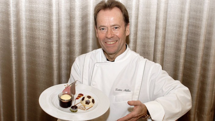 Dieter Müller blickt in Kochjacke präsentiert ein Dessert