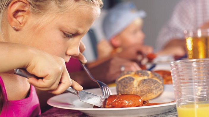 Kind isst mit Messer und Gabel eine Currywurst.