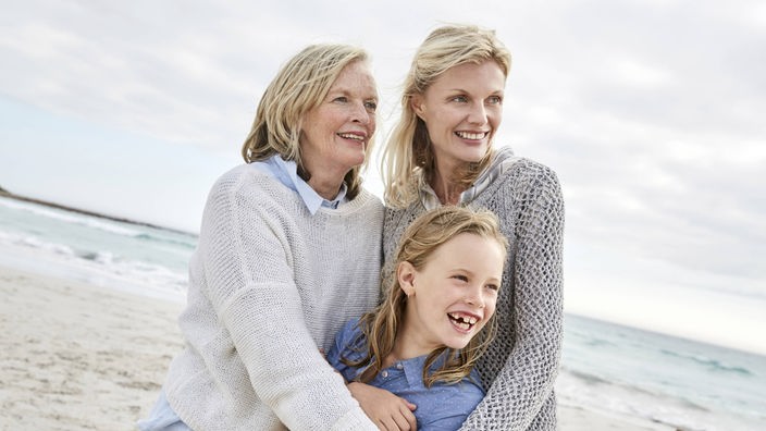Mutter, Tochter und Großmutter stehen Arm in Arm an einem Strand