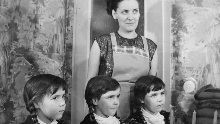 Eine Frau in Schürze und mit Hocksteck-Frisur präsentiert auf dieser Schwarzweiß-Fotografie stolz ihre Drillingsmädchen. Alle drei tragen Zöpfe, weiße Rüschenblusen und identische Strickjacken.