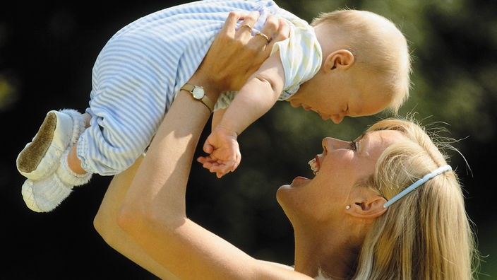 Eine Frau auf einer Wiese, die spielerisch ihr Baby hoch über ihren Kopf hält; beide lachen sich vergnügt an.