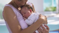 Eine junge Frau im rosa Top hält ihren schlafenden Säugling im Arm und kuschelt liebevoll mit ihm.