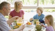 Das Bild zeigt eine fröhliche Familie mit zwei Kindern bei einer Unterhaltung am appetitlich gedeckten Frühstückstisch.
