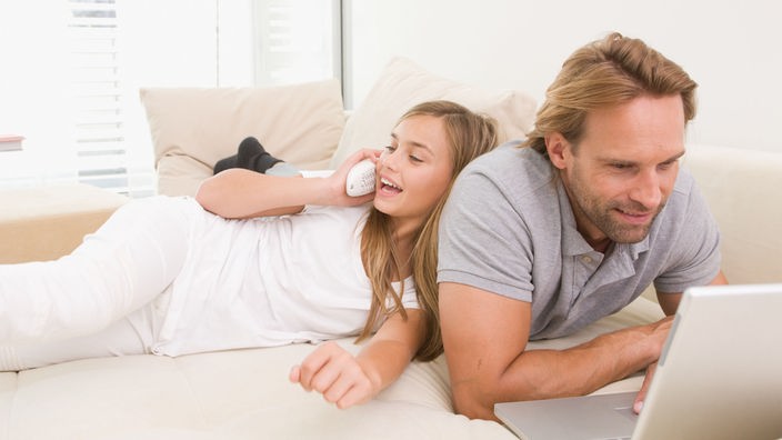 Ein Vater liegt mit seiner Tochter auf dem Sofa. Das Kind telefoniert, er arbeitet an seinem Laptop