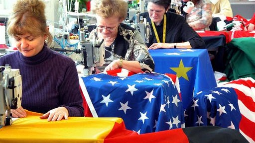 Blick in eine Flaggenfabrik. Fünf Näherinnen sitzen hintereinander an ihren Nähmaschinen. Die ersten drei Damen arbeiten jeweils an einer Deutschland-, USA- und Europaflagge.