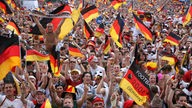 Zahlreiche Fußballfans verfolgen in einer Fanmeile ein Fußballspiel auf einer Leinwand. Die Farben 'Schwarz-Rot-Gold' findet man als Fahne, auf der Kleidung oder als 'Kriegsbemalung'.