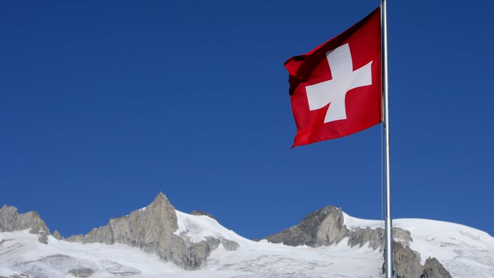 Vor einem Alpenpanorama schwenken mehrere Männer auf Stangen die quadratische Schweizer Flagge mit dem weißen Kreuz auf roten Grund.