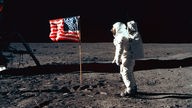 Ein Astronaut steht auf dem Mond neben der US-Flagge. Sie ist ziemlich verknittert und sieht beinahe so aus, als würde sie im Wind flattern.
