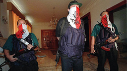 Drei bewaffnete Rebellen stehen nebeneinander, ihre Gesichter sind mit rot-weißen Tüchern maskiert.