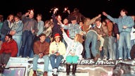 Eine Gruppe Menschen sitzt und steht mit lachenden Gesichtern auf der mit Graffiti besprühten Berliner Mauer.