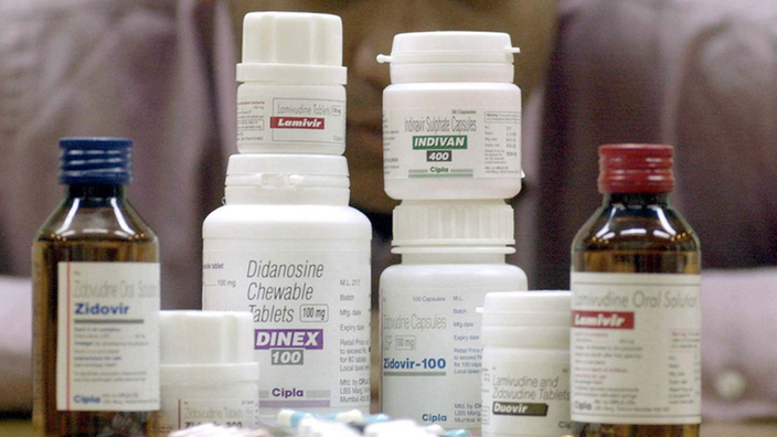Aus dem Bildhintergrund schaut ein Mann auf mehrere Dosen, Flaschen und Pillenpackungen mit Aids-Medikamenten, die im Vordergrund übereinander liegen und stehen.