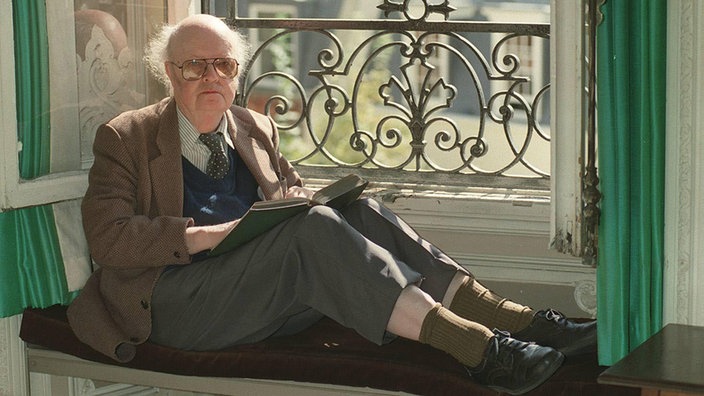 John Bayley sitzt vor einem Fenster und hält ein Buch in der Hand.