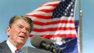 Ronald Reagan während einer Rede am Mikrophon, im Hintergrund die Amerikanische Flagge.