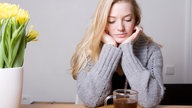 Eine junge Frau sitzt am Tisch und schaut traurig auf ihre Teetasse.