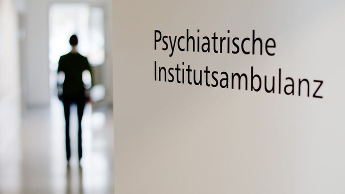 Das Bild zeigt das Hinweisschild der psychiatrischen Abteilung einer Klinik. Im Hintergrund steht ein Mensch im Flur.