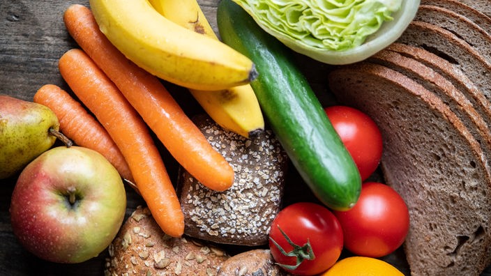 Verschiedene gesunde Lebensmittel auf einem Tisch: Vollkornbrot, Salat, Bananen, Möhren, Gurke und anderes Obst und Gemüse