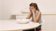 Eine Frau sitzt am Esstisch vor einem großen leeren Teller und schlägt die Hände vors Gesicht