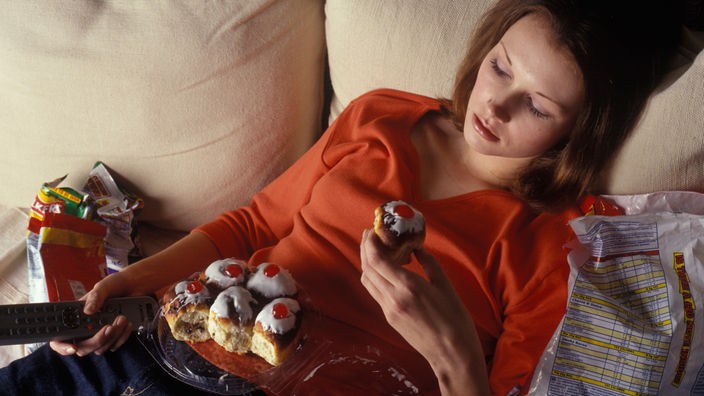 Eine Frau auf dem Sofa mit vielen Kuchenstücken und Chipstüten