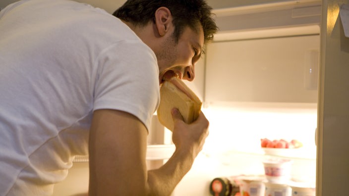 Ein Mann steht vor einem geöffneten Kühlschrank und isst ein Sandwich