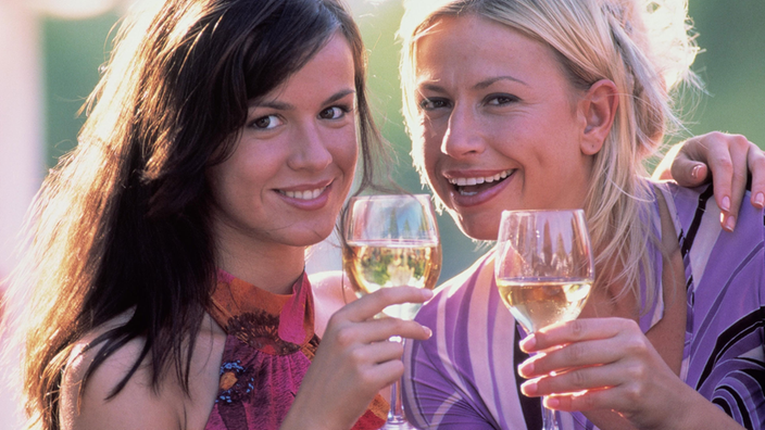 Zwei Frauen halten jeweils ein Glas mit Weißwein. Sie lächeln in die Kamera.