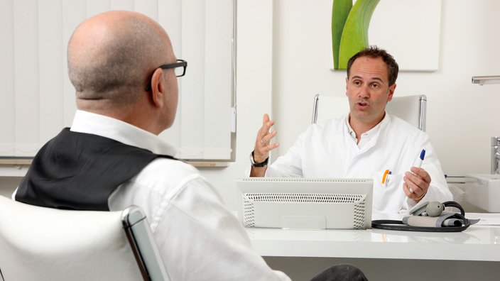 Ein Mann, der mit dem Rücken zur Kamera sitzt, spricht mit seinem Arzt, der gegenüber von ihm am Schreibtisch sitzt. | Bildquelle: imageBROKER / Jochen Tack Ein Mann, der mit dem Rücken zur Kamera sitzt, spricht mit seinem Arzt, der gegenüber von ihm am Schreibtisch sitzt.