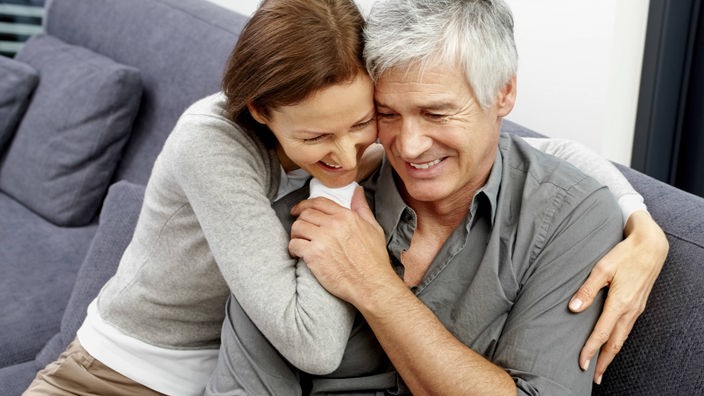 Mann und Frau sitzen auf einem grauen Sofa. Sie umarmt ihn von der Seite, beide lächeln.