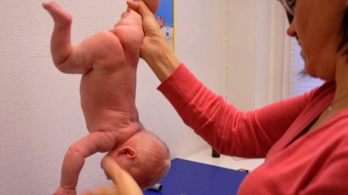 Eine Ärztin hält ein Baby an einem Bein in die Luft und lässt es kopfüber über einer Ablage baumeln.