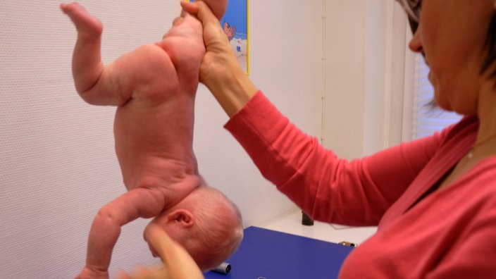 Eine Ärztin hält ein Baby an einem Bein in die Luft und lässt es kopfüber über einer Ablage baumeln. | Bildquelle: WDR/Klaus Rose Eine Ärztin hält ein Baby an einem Bein in die Luft und lässt es kopfüber über einer Ablage baumeln.