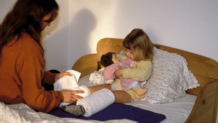 Ein Mädchen sitzt auf einem Bett, seine Mutter links daneben. Es trägt Wickel um die Waden und ist augenscheinlich krank.
