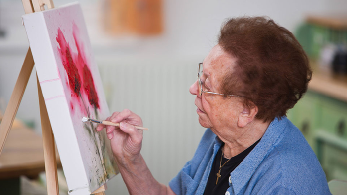Eine alte Frau sitzt vor einer Staffelei und malt.