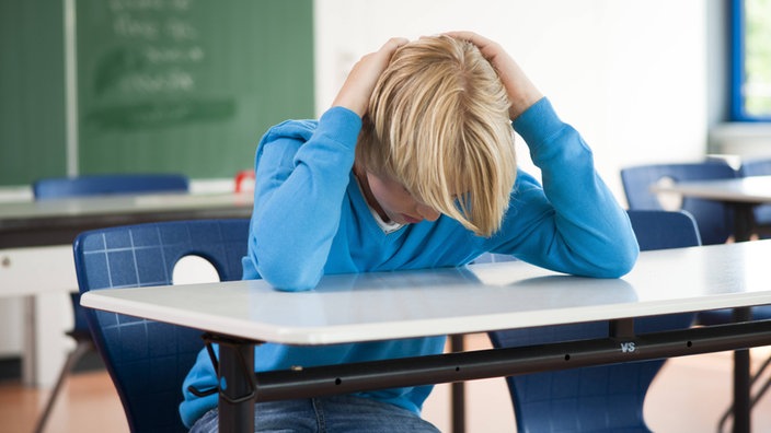 Blonder Junge sitzt in einem Klassenzimmer. Er hat die Arme und Hände über seinem Kopf zusammen geschlagen. | Bildquelle: WDR/imago stock&people Blonder Junge sitzt in einem Klassenzimmer. Er hat die Arme und Hände über seinem Kopf zusammen geschlagen.