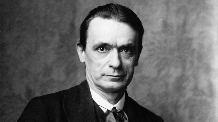 Schwarzweiß-Foto von Rudolf Steiner in einem Lehnstuhl sitzend mit Brille in der Hand.