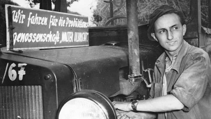 Schwarzweißaufnahme: Ein freiwilliger Helfer in der LPD 'Walter Ulbricht' in Merxleben steht neben einem Traktor (1952).
