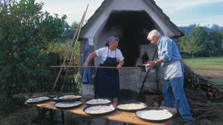 Eine älteres Paar steht mit mehreren Blechen Flammkuchen vor einem Backhäuschen.