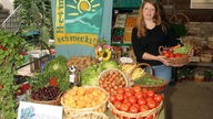 Verkäuferin in einem Hofladen trägt einen Korb frischen Gemüses neben einem Werbetransparent mit dem Schriftzug 'Heimat schmeckt'.