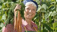 Eine ältere Frau hält lächelnd einen Bund Karotten in die Kamera.