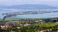 Eine Luftaufnahme des Bodensees, umgeben von grünen Wiesen und kleinen Häusern mit roten Dächern. In der Bildmitte die Insel Reichenau.