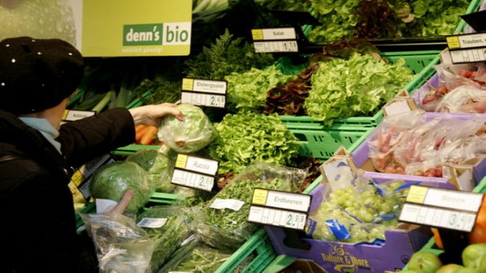 Eine Frau haelt in einem Supermarkt Bio-Salat in der Hand, sie steht beim Bio-Gemüse.