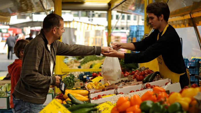 Auf einem Wochenmarkt bezahlt ein Mann bei einem jüngeren am Obst und Gemüsestand