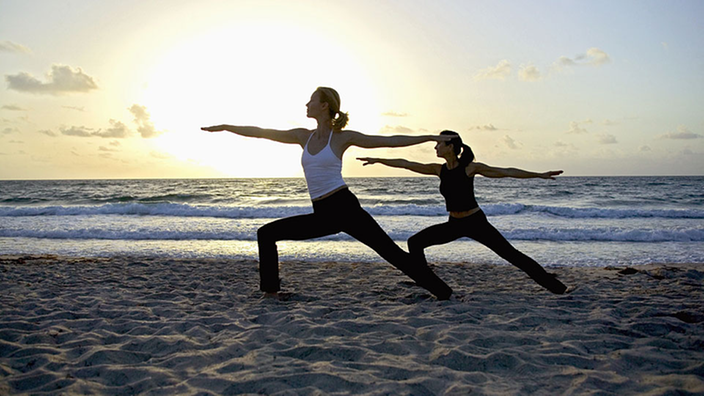 Zwei Frauen praktizieren Yoga am Sandstrand bei untergehender Sonne.