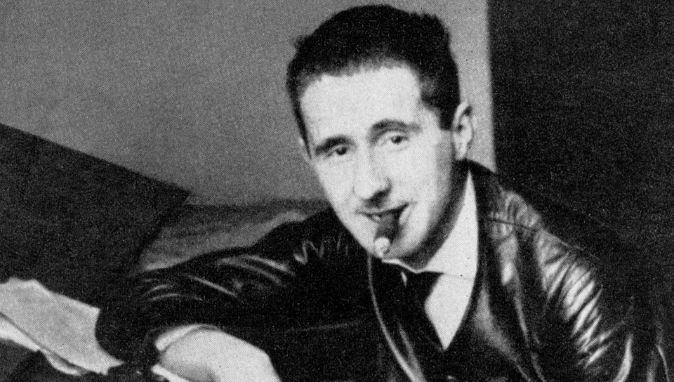 Portrait von Bertolt Brecht in Lederjacke, Zigarre rauchend an ein Klavier gelehnt.