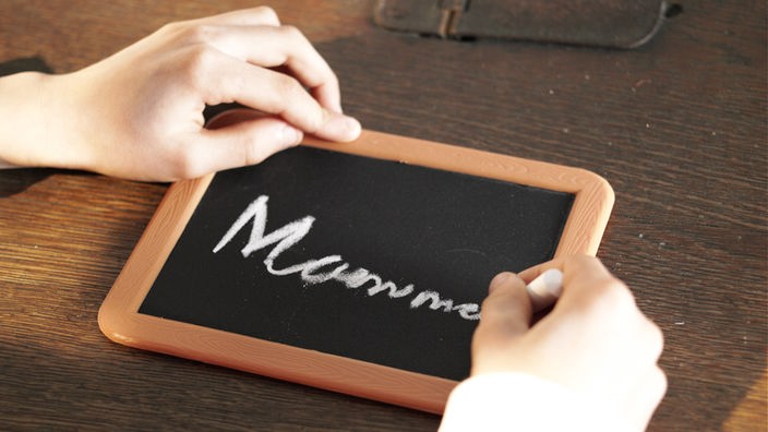 Auf einer kleinen Tafel ist das Wort "Mamma" zu lesen. Davor befinden sich zwei Kinderhände, die rechte hält ein Stück Kreide