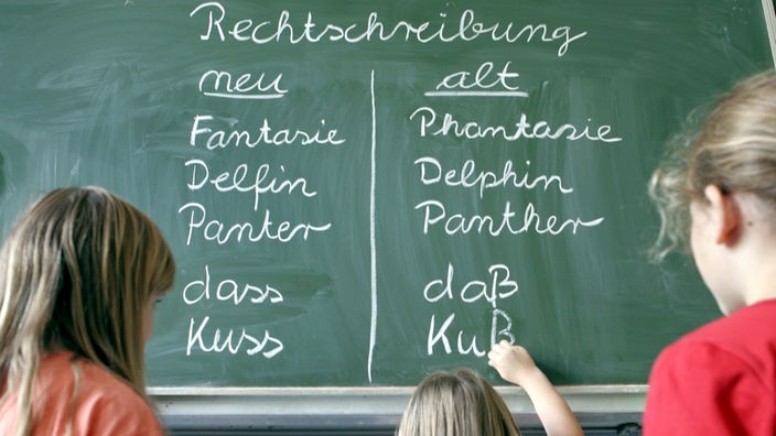 Drei Mädchen schreiben zum Vergleich Wörter in der neuen und alten Rechtschreibung an eine Tafel