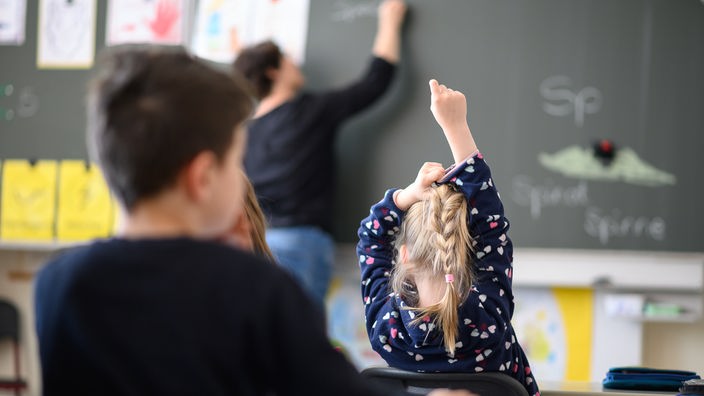 Ein Mädchen in einer Schulklasse streckt den Finger nach oben, während eine Lehrerin etwas an die Tafel schreibt