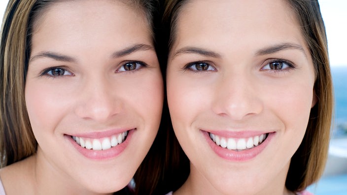 Das Foto zweigt zwei Teenagerinnern, die Zwillinge sind. Sie lächeln freundlich in die Kamera.