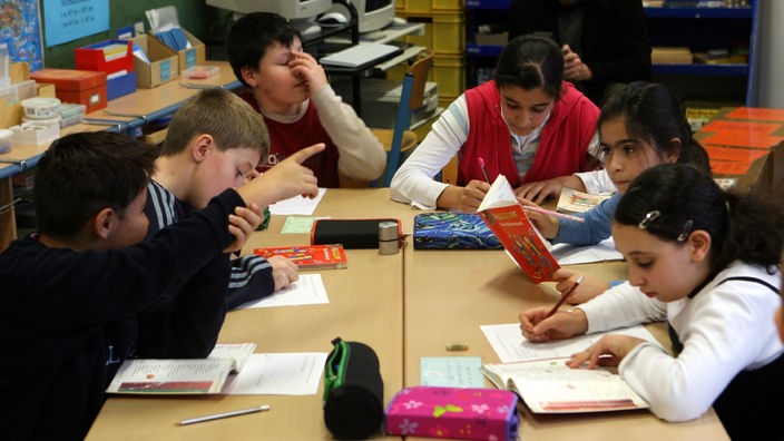 Schüler einer Grundschulklasse sitzen sich an Tischen gegenüber und lösen Aufgaben aus einem Buch.