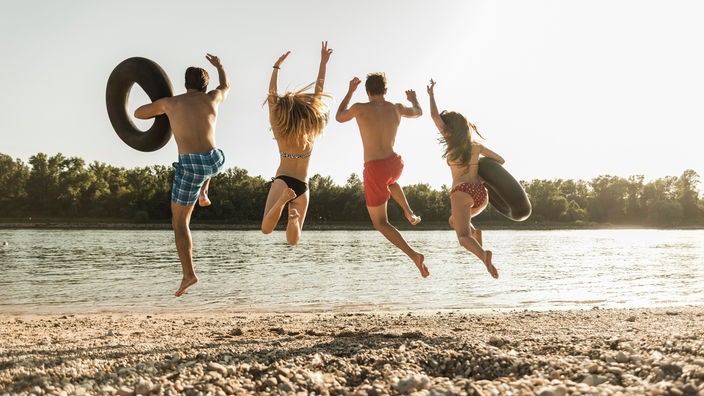 Vier junge Menschen, zwei Jungs, zwei Mädchen, springen mit Schwimmreifen in einen See. Sie haben der Kamera den Rücken zugewendet.
